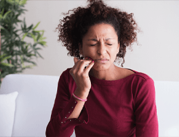 A woman wearing a maroon blouse and holding her cheek in pain because of escalating gum disease