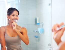 A young woman wearing a gray tank top and brushing her teeth while in the bathroom