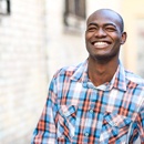 A young man standing outside in a plaid shirt smiling after receiving a CEREC dental crown