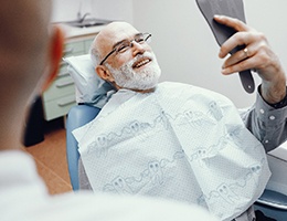 An older man with glasses looking at his new smile in the mirror while seated in the dentist’s chair