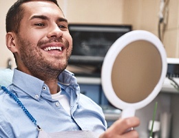 A young male wearing a blue button-down shirt and looking at his new smile in the mirror at the dentist’s office